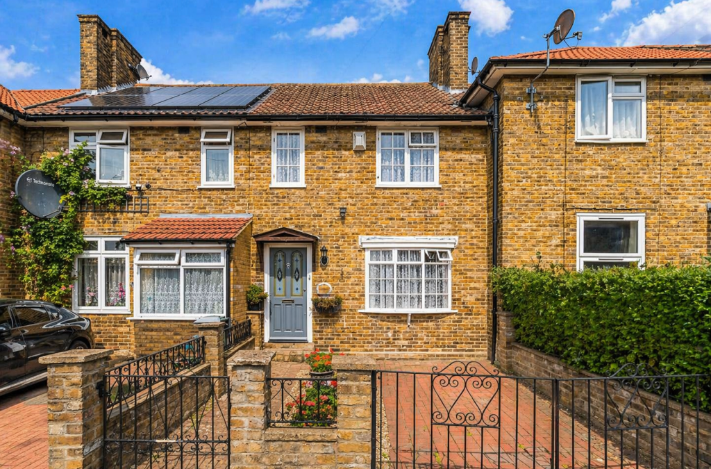 Brick terraced house with blue door, small front garden, and ornate iron gate; brick driveway and neighboring homes under a blue sky.