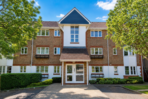 Three-story brick apartment building with a central entrance under a gabled blue-trimmed peak, white accents, and greenery around.