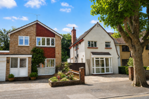 Two houses on a sunny suburban street with a large tree on the right and small front gardens.