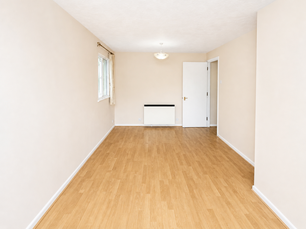 Empty, neutrally colored room: beige walls, light wood laminate flooring, window with cream curtain on the left, and a white door on the far wall.