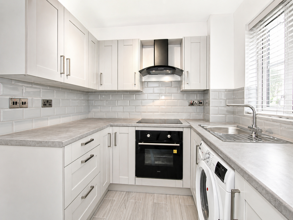 Bright, modern white kitchen with shaker cabinets, grey countertops, and a stainless-steel range hood over a built-in oven and stovetop far wall.