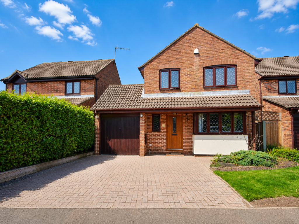 Brick two-story house with brown tiled roof, attached garage, wooden front door, and a paved driveway, under a blue sky with a hedge on the left.