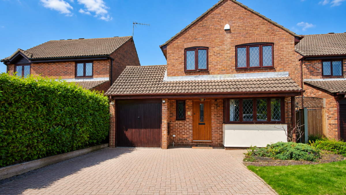 Brick two-story suburban house with attached garage and a paved driveway under a blue sky with a hedge on the left.
