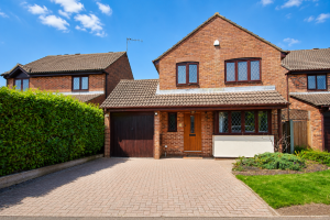 Brick two-story suburban house with attached garage and a paved driveway under a blue sky with a hedge on the left.