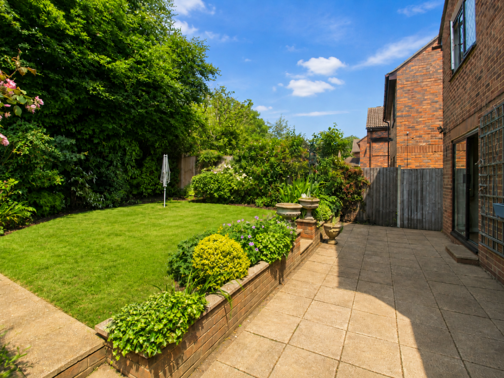 Sunlit backyard with a manicured lawn, raised brick planter filled with flowers, and a tiled patio beside a brick house on the right.