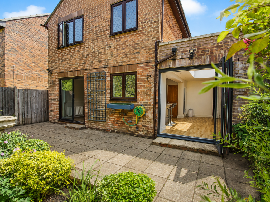 Back of a brick house with a paved patio; large glass doors open to a bright kitchen area, a garden hose reel mounted on the wall, and a trellis beside a window.