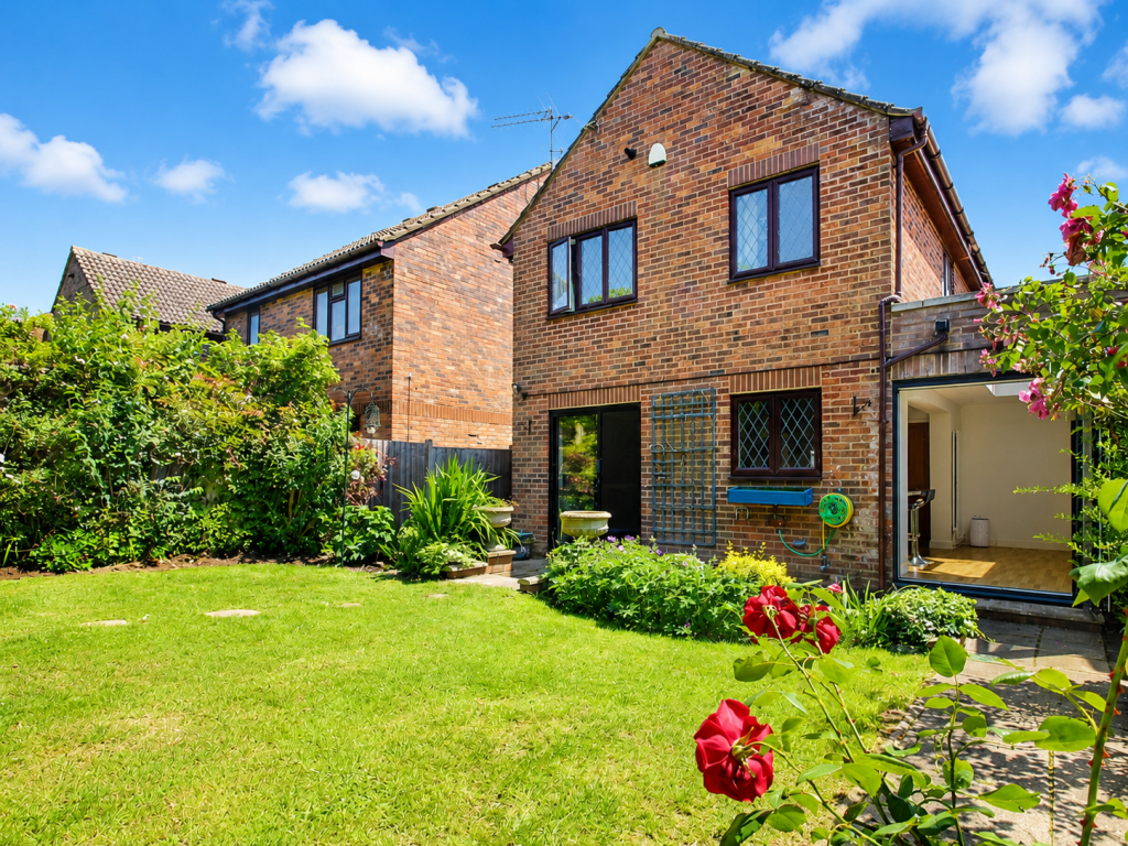 Brick two-story house with a well-kept lawn and bright blue sky, red roses in the foreground, and garden plants along the edge of the yard.