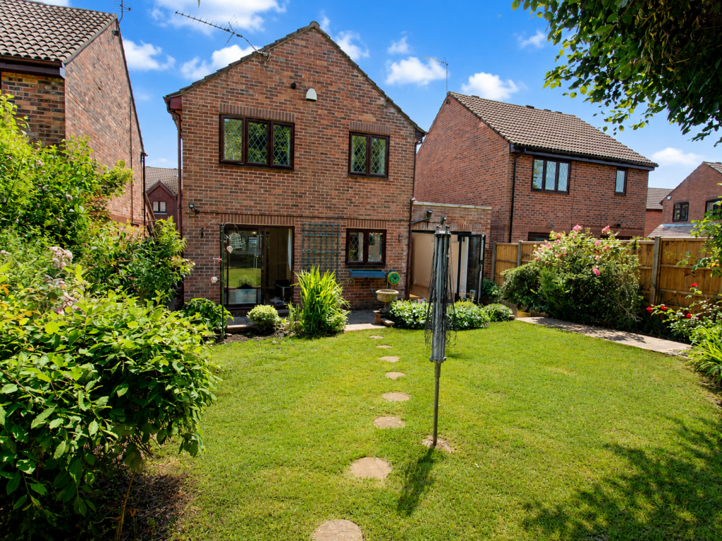 Backyard of brick houses with a green lawn, stepping-stone path, and garden beds along a wooden fence under a blue sky.