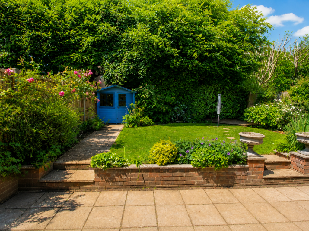 Blue wooden shed at the end of a stone path in a lush backyard with green hedges and pink flowers nearby.