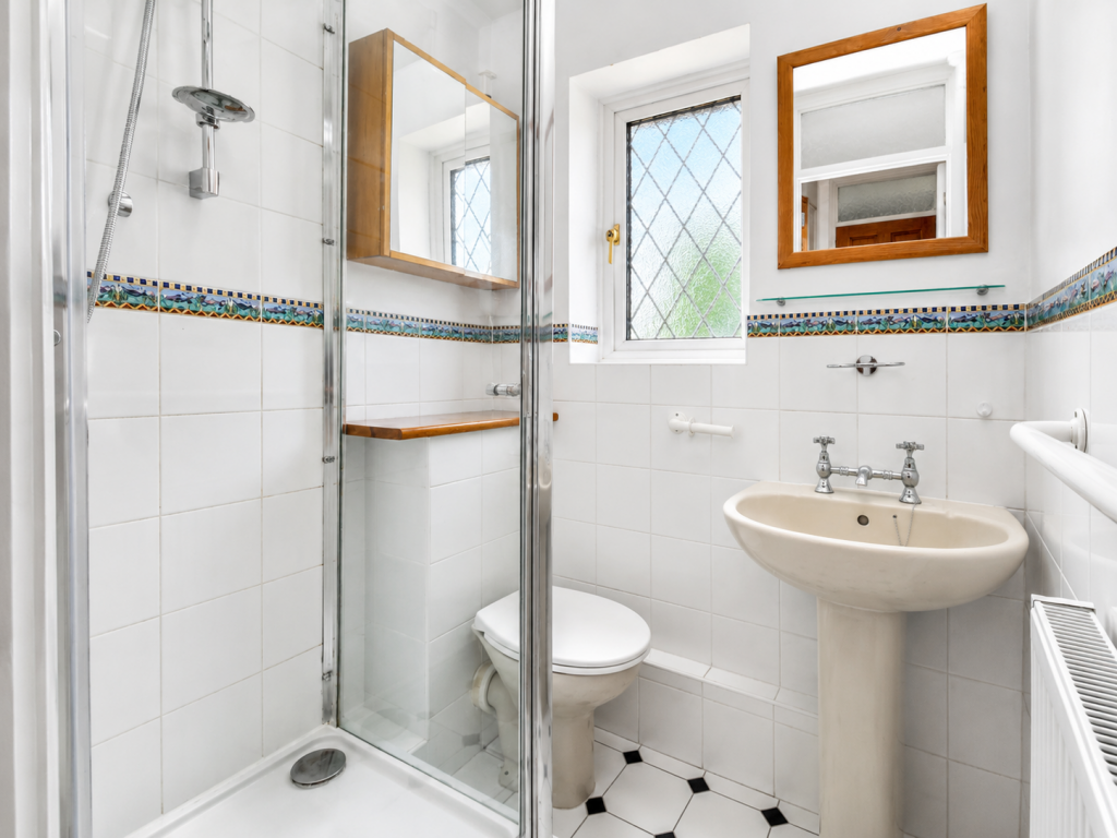 Small tiled bathroom with a glass shower enclosure, pedestal sink, and toilet; decorative mosaic border along the wall and a frosted window above the sink mirror.