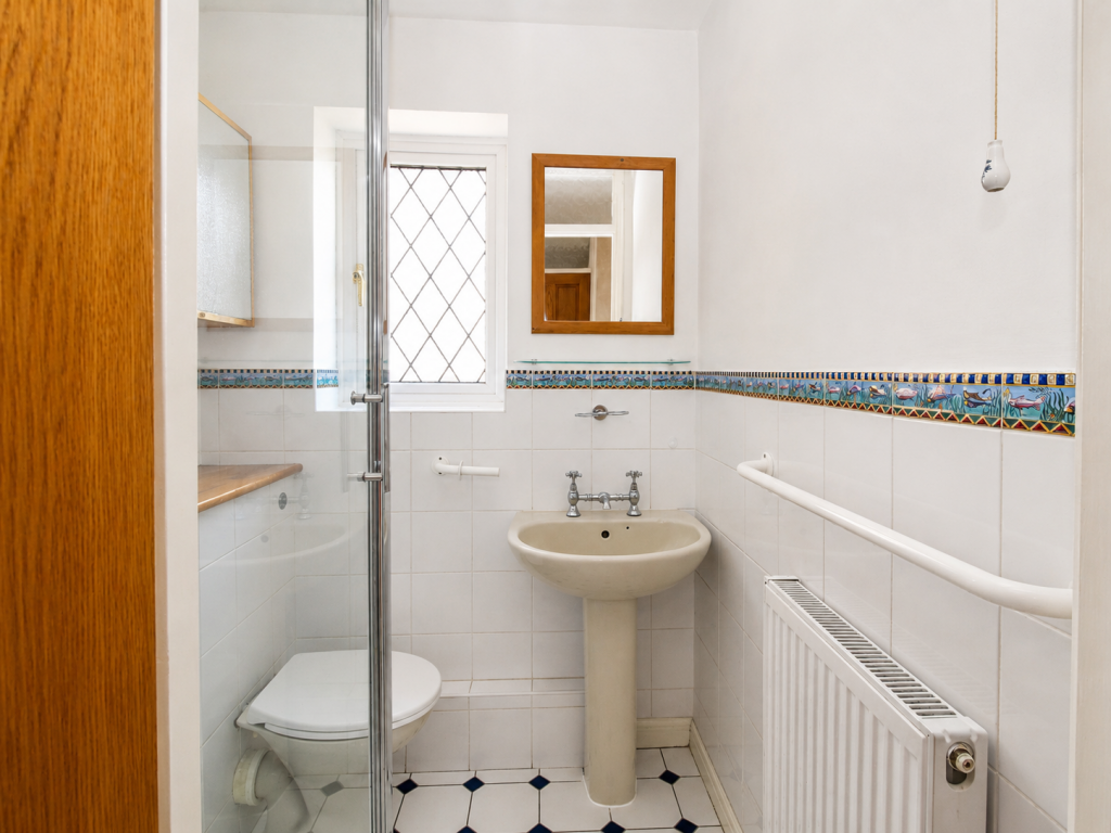 Small bathroom with pedestal sink, toilet, and glass shower enclosure; decorative tile border and a wooden-framed mirror with window in the background.