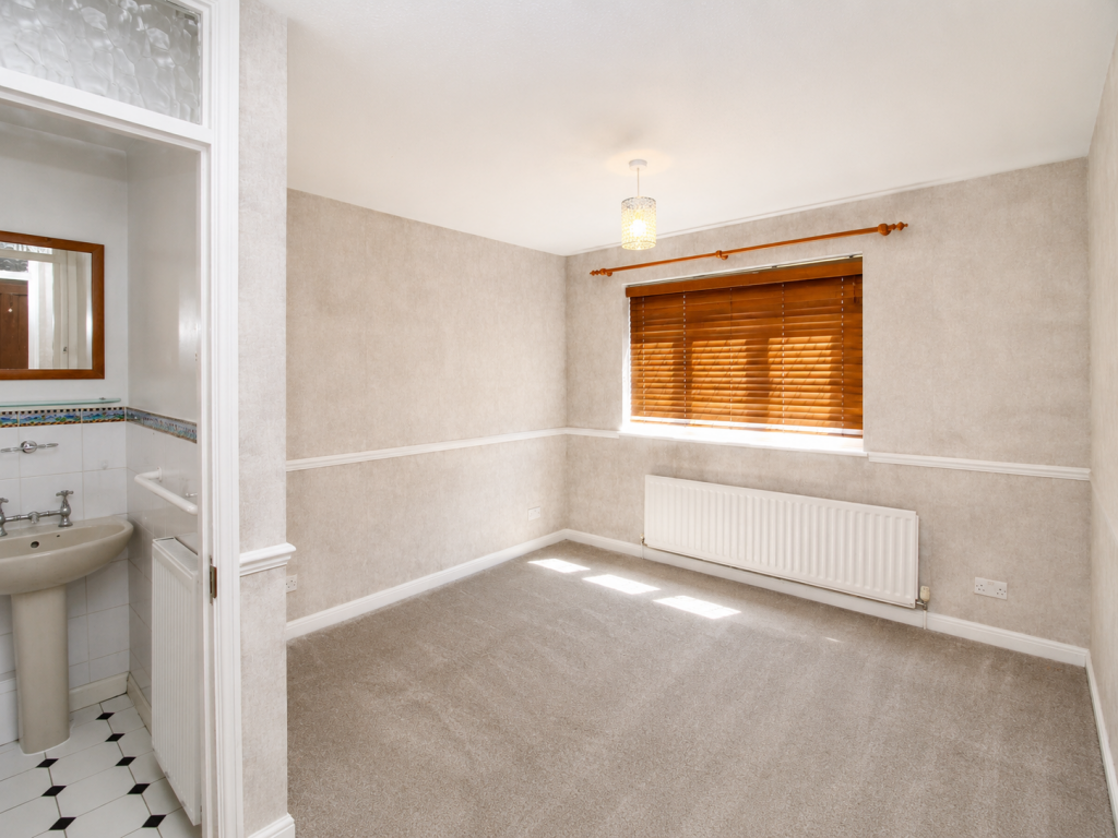 Empty beige living room with a window covered by wooden blinds, radiator along the wall, and a doorway to a small bathroom with a pedestal sink visible on the left.