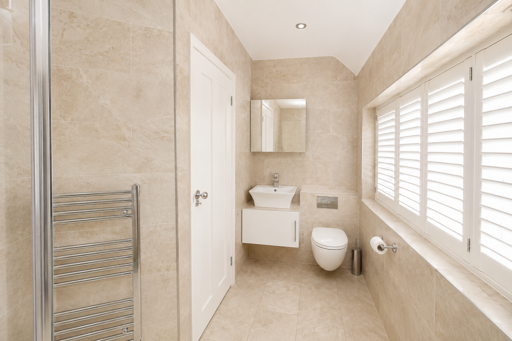 Beige-tiled bathroom with a wall-mounted sink, mirror, and wall-hung toilet next to a window with white shutters.