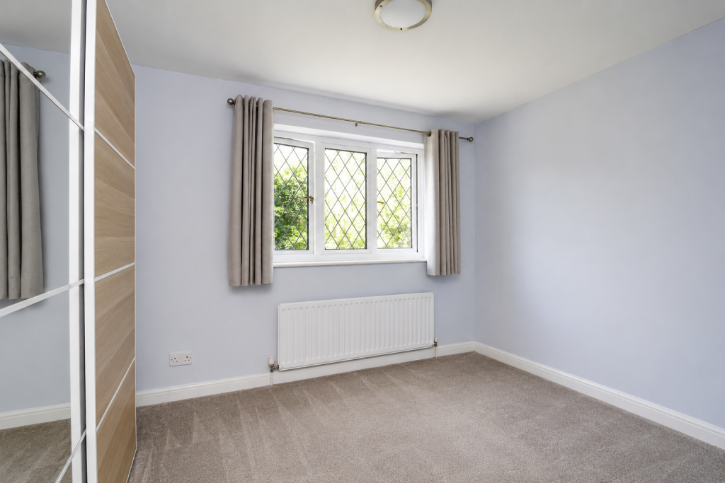 Empty light-blue bedroom with a large window featuring diamond-lead panes, beige curtains, and a white radiator beneath the window. A mirrored wardrobe and beige carpet complete the space.