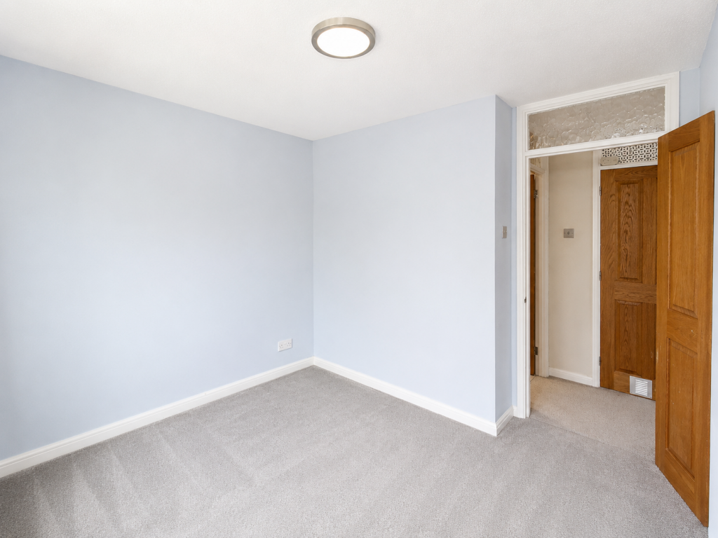 Empty small room with light blue walls, beige carpet, and a circular ceiling light; a wooden door is open to a hallway.