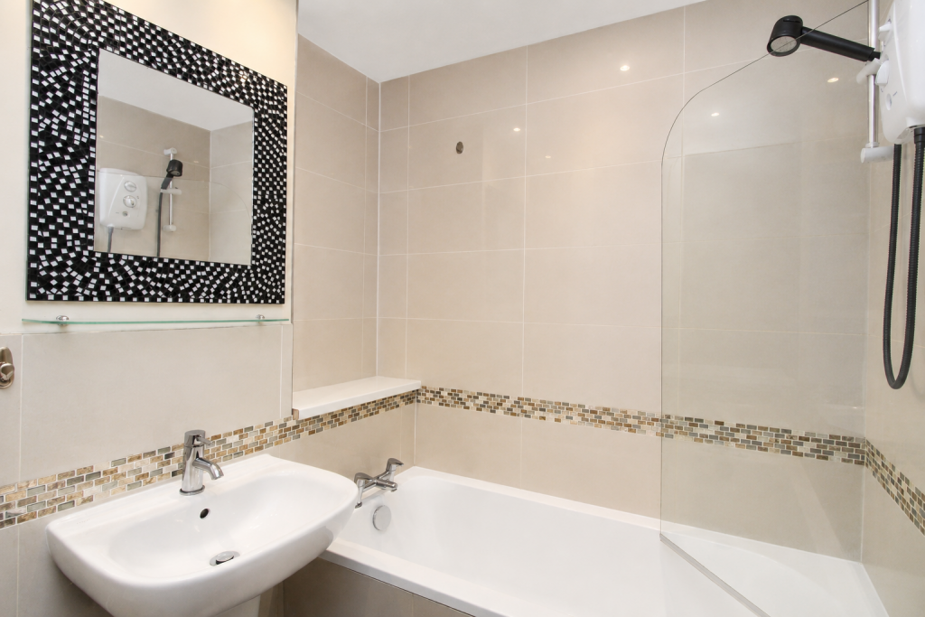Modern bathroom with beige tiled walls, a white sink, and a bathtub; a black-and-white mosaic-framed mirror above a glass shelf, and a shower head on the right wall.