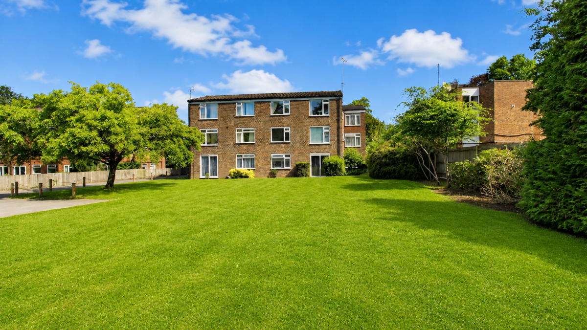 Three-story brick apartment building facing a wide green lawn under a bright blue sky with scattered clouds.