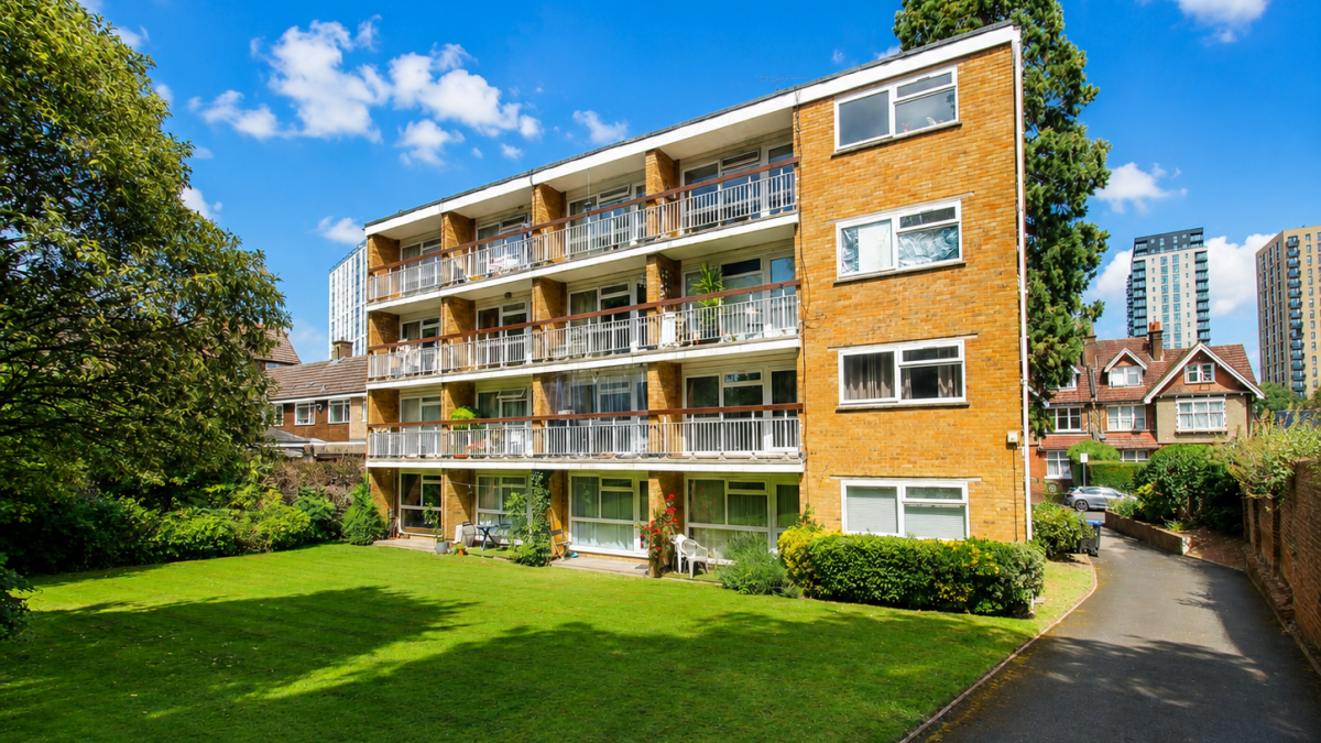 Brick apartment building with four stories and balconies, set in a green lawn under a bright blue sky.