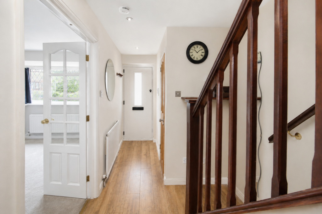 Bright hallway with a dark wooden staircase on the right, white walls, and a round clock above the wall switch, leading to a front door at the end.