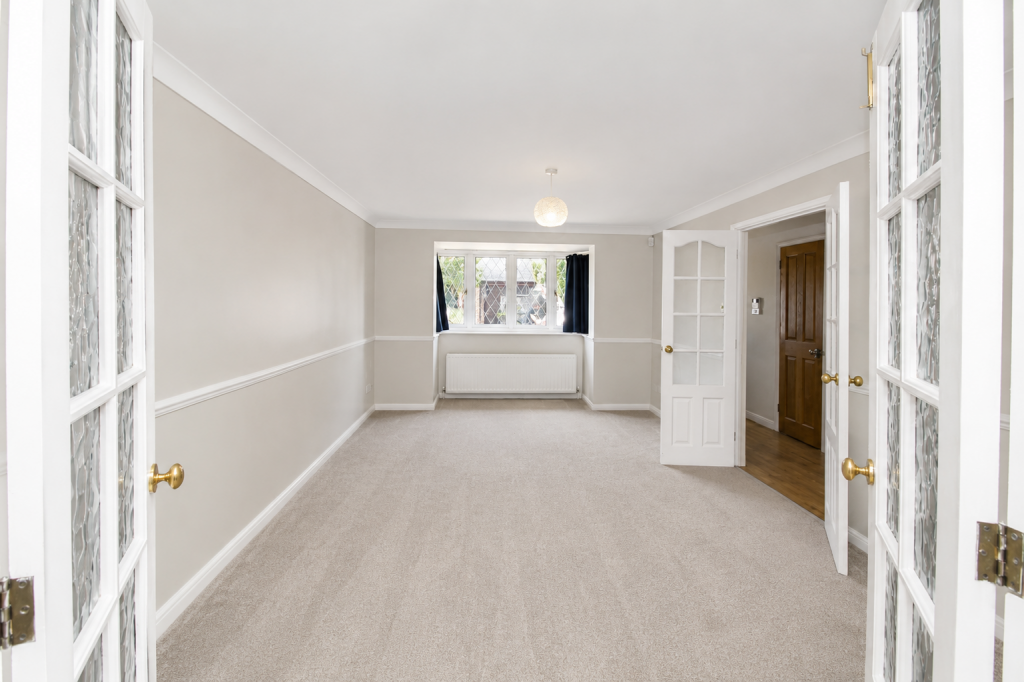 Bright empty living room with beige walls, white trim, and light carpeting; a window with a radiator beneath it.