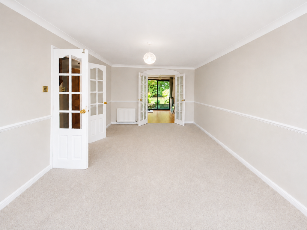 Bright empty living room with beige walls, white trim, and carpet; glass-paned French doors open to a sunlit garden.
