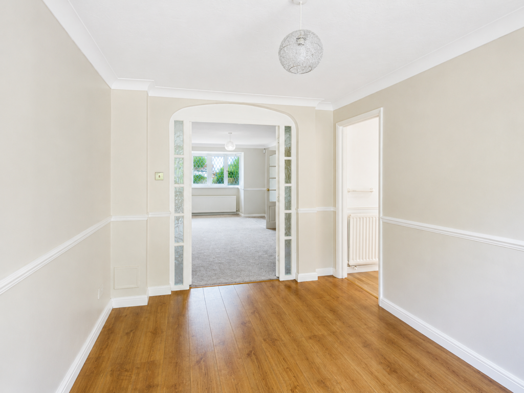 A hallway with light walls and wooden floor leading to a room with windows and a hanging light fixture; glass-panel doors are open between spaces