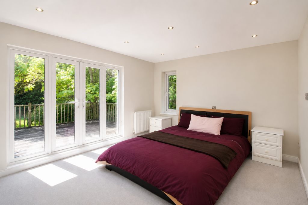 Bright bedroom with large glass doors opening to a wooden deck and lush green trees; queen bed with burgundy bedding and white nightstands on each side.