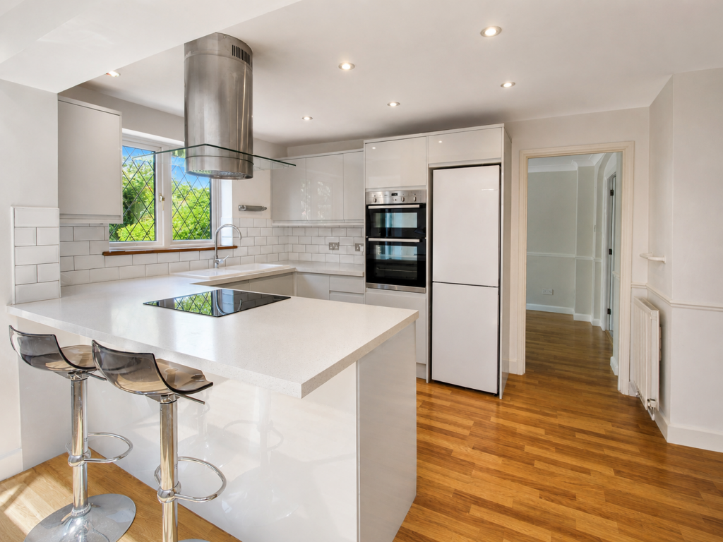 Bright white kitchen with an island, induction stove, stainless appliances, and wood flooring visible near a window with greenery outside.