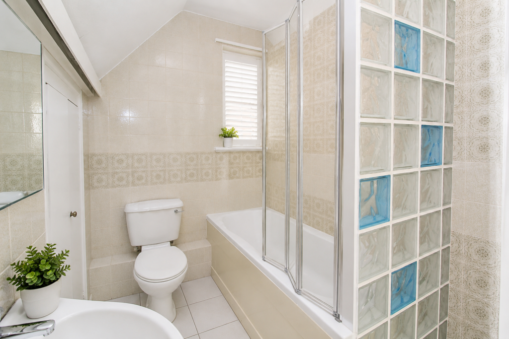 Bright beige tiled bathroom with a toilet, corner tub with sliding glass doors, and a glass block partition.