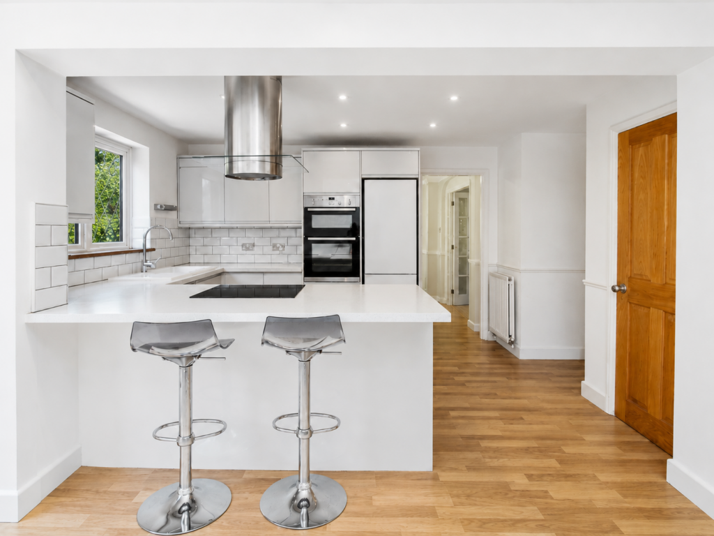 Bright, modern white kitchen with an island, two chrome bar stools, and stainless-steel appliances; warm wood floor and a window to the left.