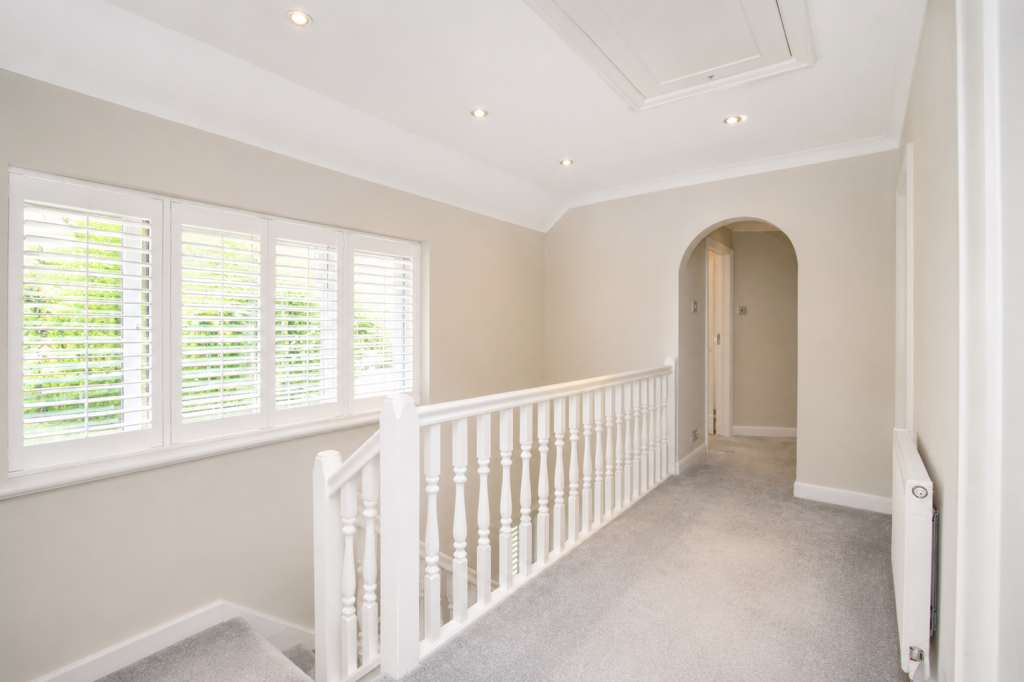 Bright upstairs hallway with a white balustrade, beige walls, and large window shutters letting in natural light on the left.