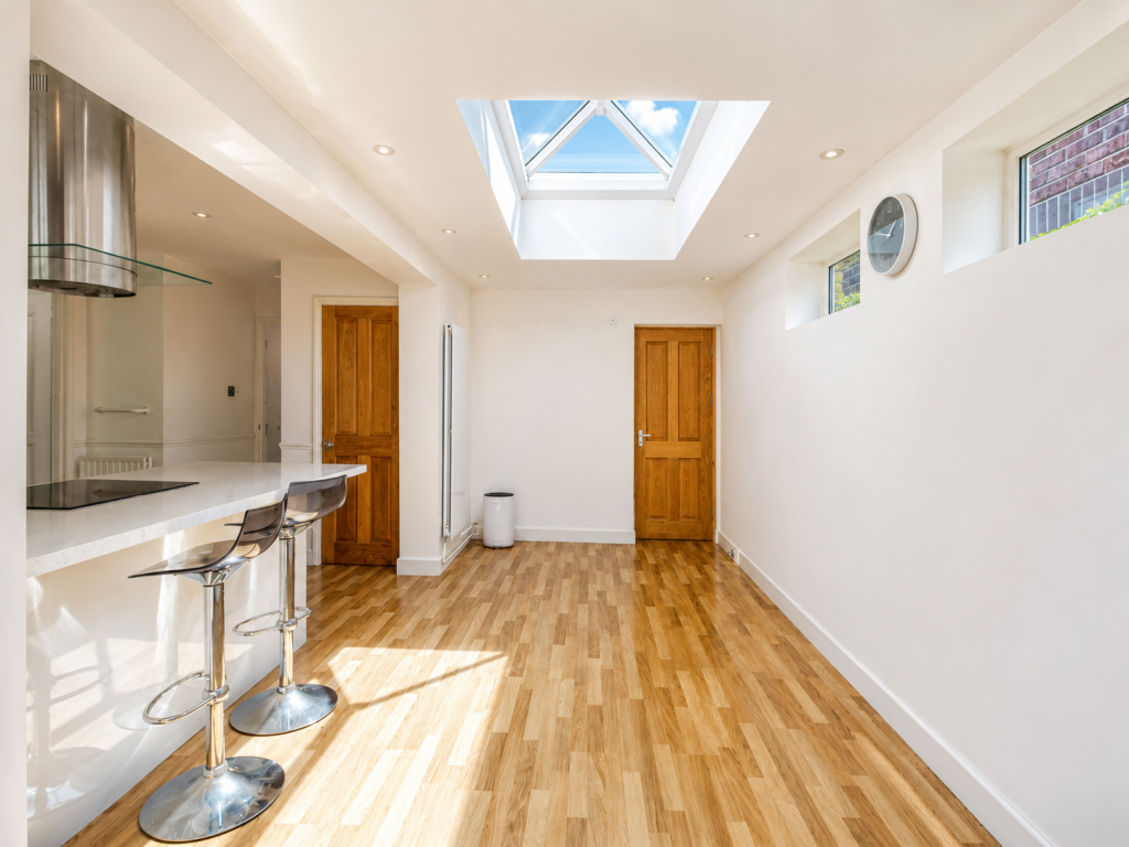 Bright open-plan kitchen with white walls, a skylight, and a wood floor; a white countertop bar with chrome stools is on the left.