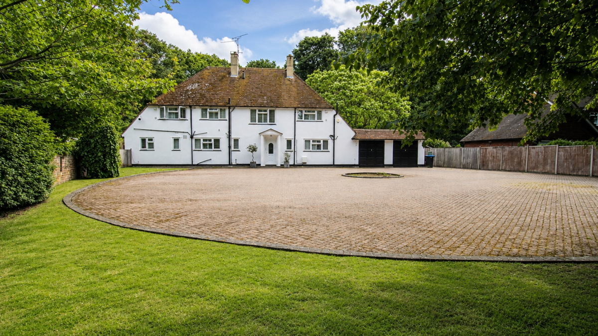 White country house with a brown tiled roof, circular gravel driveway, and green lawn with trees framing the scene.