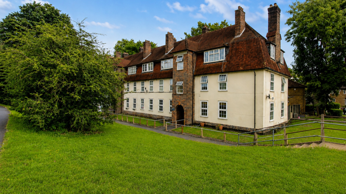 Historic cream-colored building with a steep red-tiled roof and multiple chimneys beside a green lawn under a blue sky.