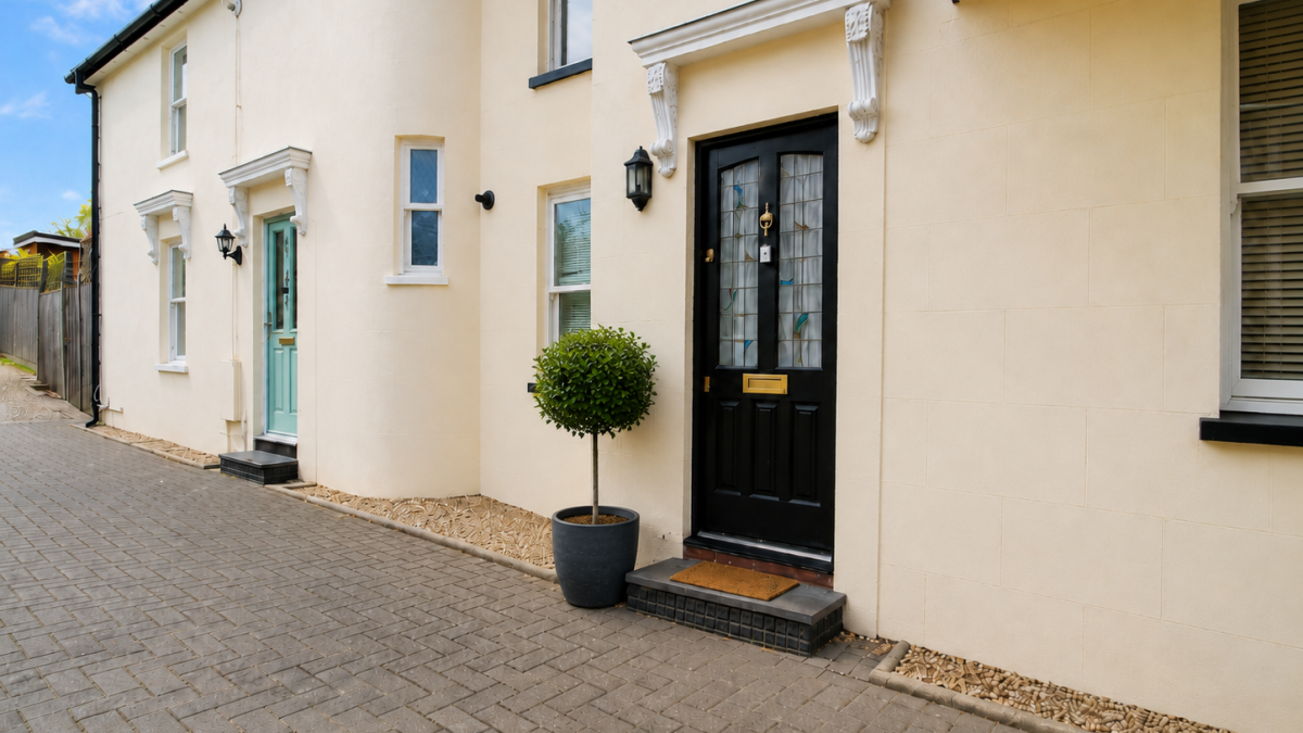 Beige townhouse façade with a black front door and a round, trimmed potted tree by the entry on a paved walkway.