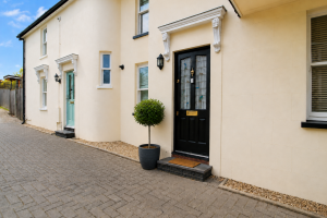 Beige townhouse façade with a black front door and a round, trimmed potted tree by the entry on a paved walkway.