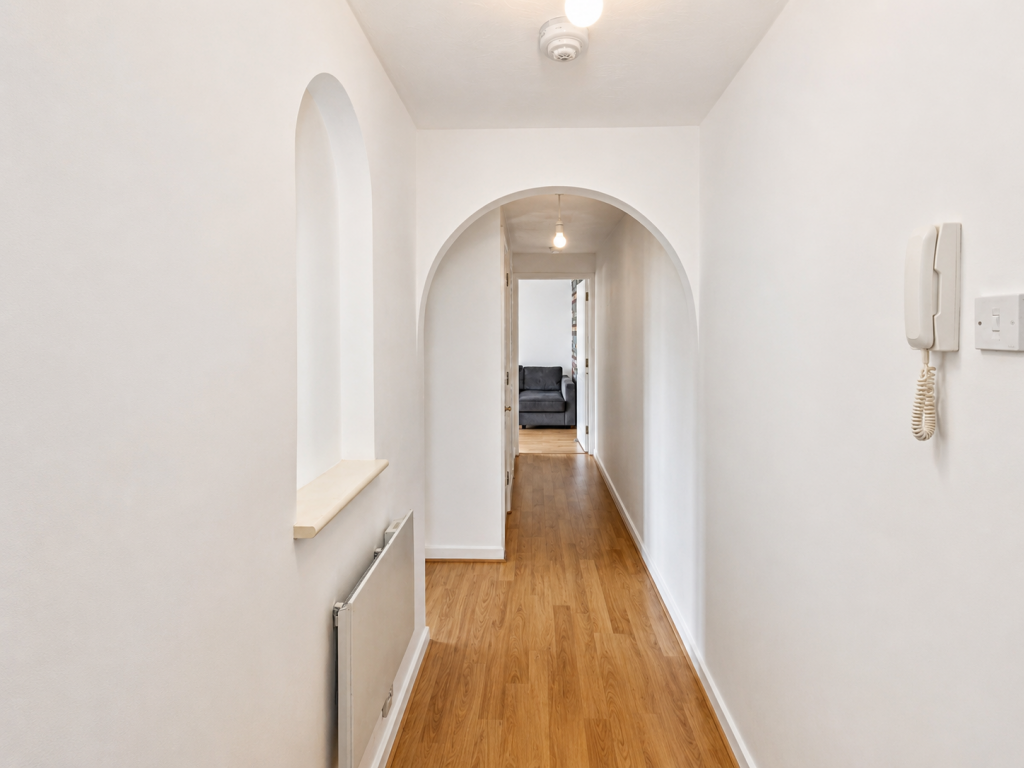 Bright white hallway with a wooden floor and an arched doorway leading to a living room with a gray sofa visible at the end, intercom on the right wall.