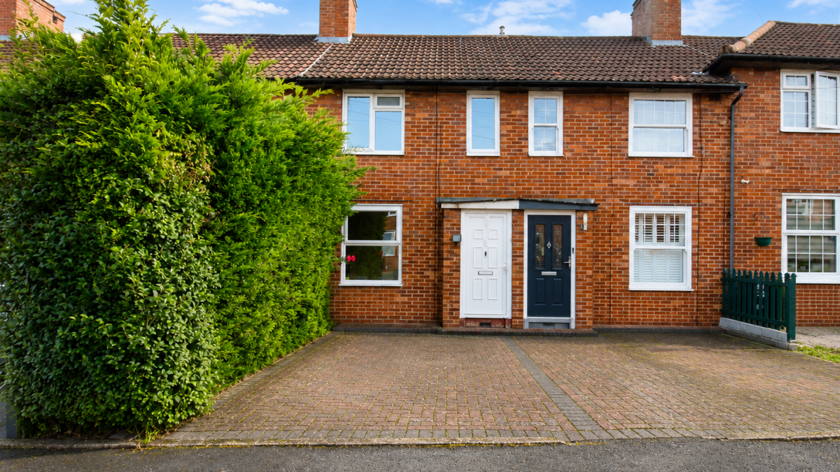 Brick terraced house with white and dark blue front doors, a tall green hedge on the left, and a brick driveway.