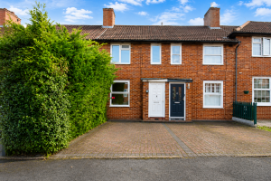 Brick terraced house with white and dark blue front doors, a tall green hedge on the left, and a brick driveway.
