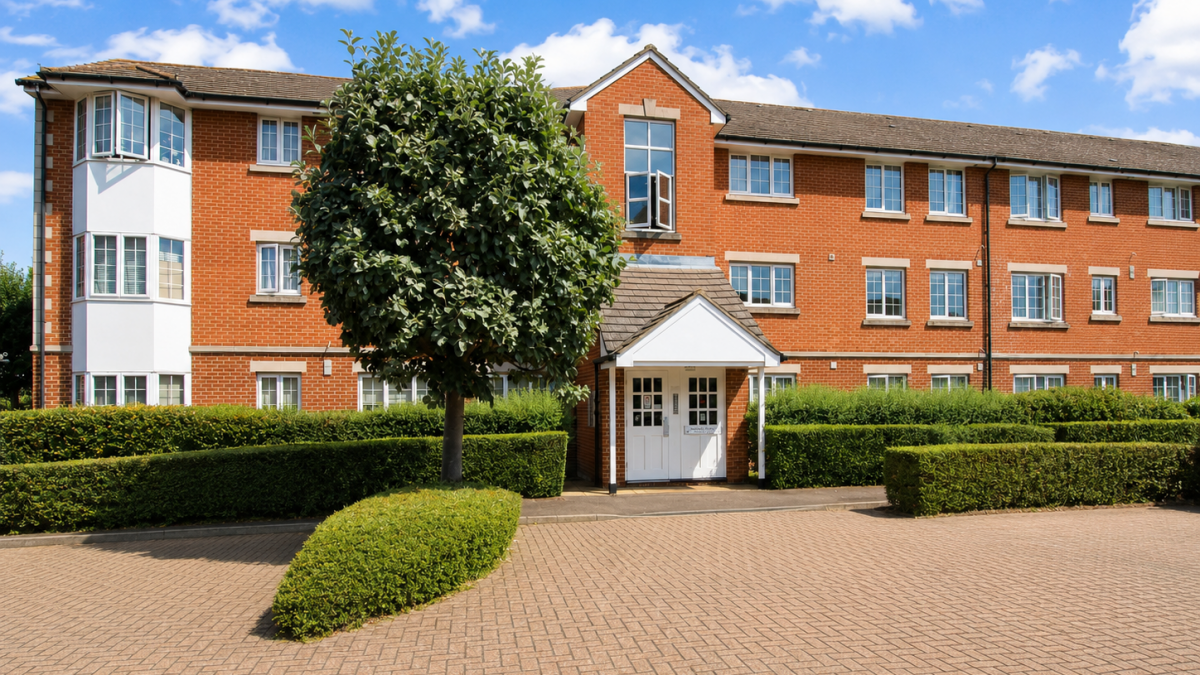 Red brick apartment building with white entrance doors, trimmed hedges, and a large tree in front on a sunny day.