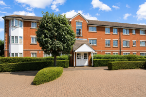 Red brick apartment building with white entrance doors, trimmed hedges, and a large tree in front on a sunny day.