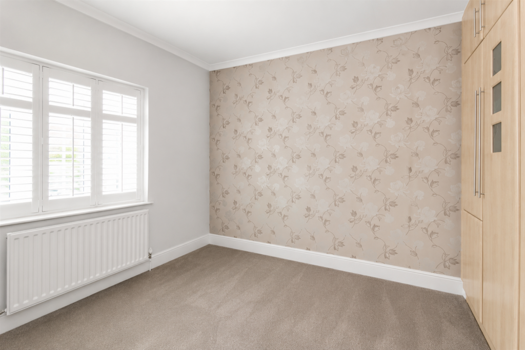 Bedroom corner with beige floral wallpaper on one wall, white window with shutters, radiator below, and built-in wooden wardrobe.