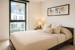 Modern bedroom with beige bed and pillows, dark wood frame, beside a large window and a nightstand with lamp.