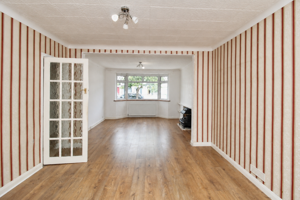 Empty living room with red-and-beige striped wallpaper, wooden floor, and a glass-panel door opening to a bright back room with a bay window.