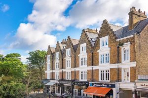 Row of brick townhouses with decorative gables along a sunny street, storefronts on the ground level and a blue sky above.